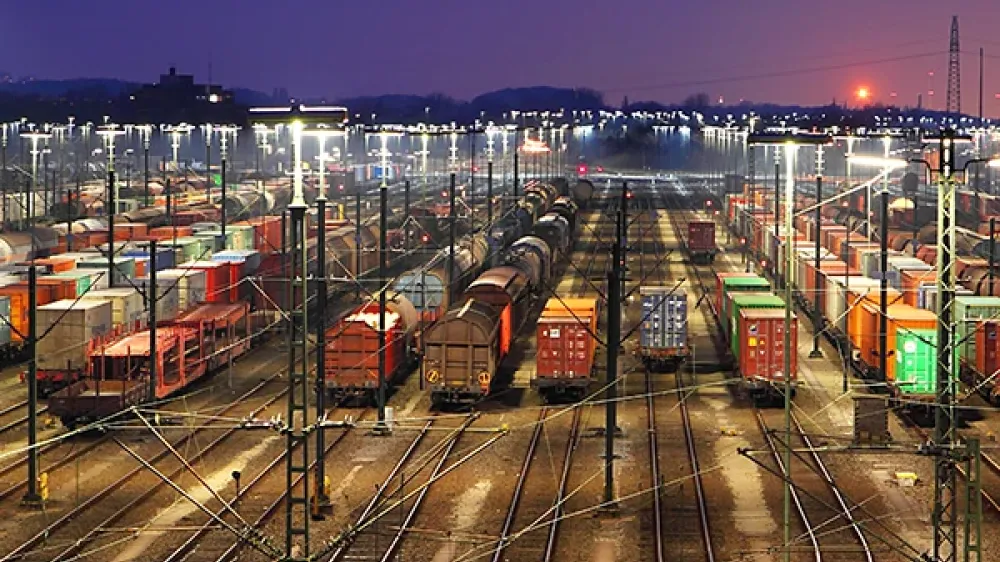 Zone de la gare avec de nombreux rails, aiguillages et lignes électriques, devant la silhouette d’une ville.