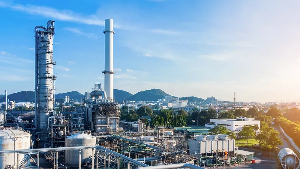 Industrial complex with pipes, storage tanks, and tall chimneys under a blue sky; mountains and a cityscape are visible in the background.