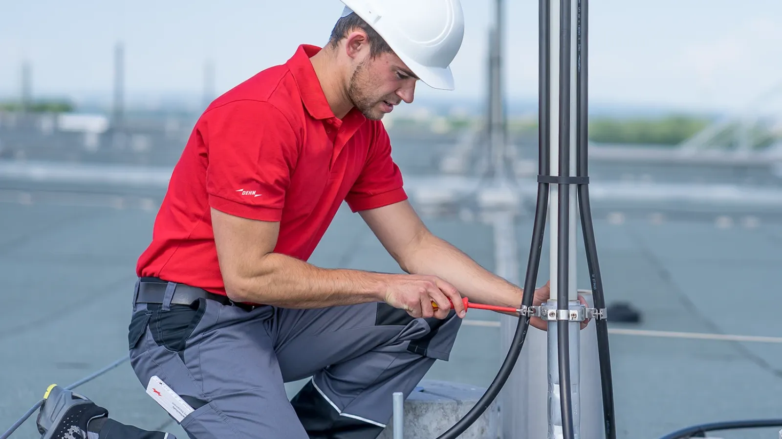 Technician in red shirt and white hard hat installing grounding cables on a rooftop lightning protection mast.