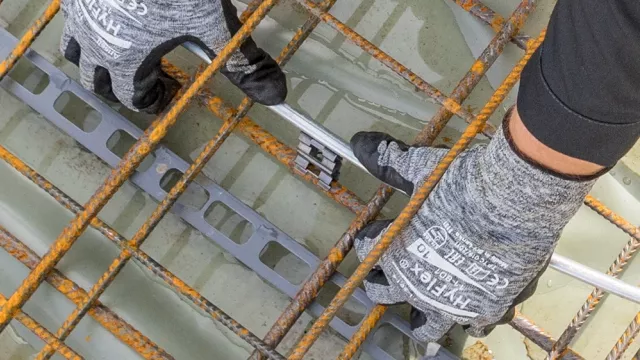 Gloved hands attach a metal connector to round steel rebars on a wet rebar grid, viewed from above.