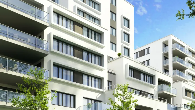 Modern white apartment buildings with multiple balconies, large windows, and surrounding green trees under a clear blue sky.