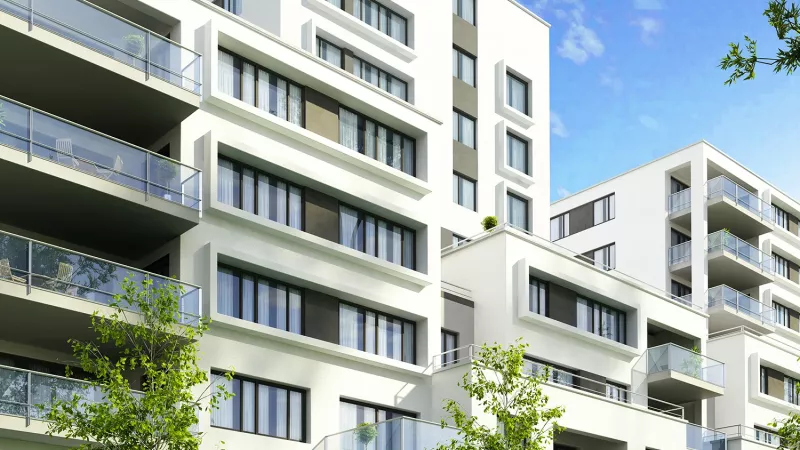 Modern white apartment buildings with multiple balconies, large windows, and surrounding green trees under a clear blue sky.