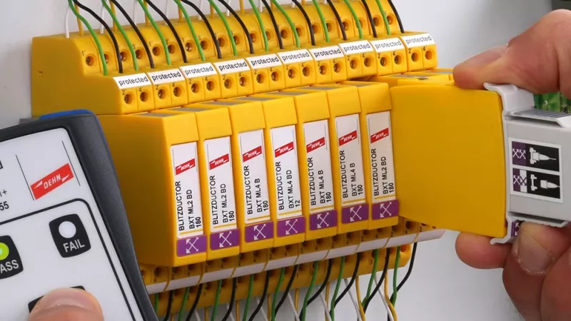 Hand holding a measuring device next to a row of yellow surge protection modules while another hand holds a removed module with test adapter.