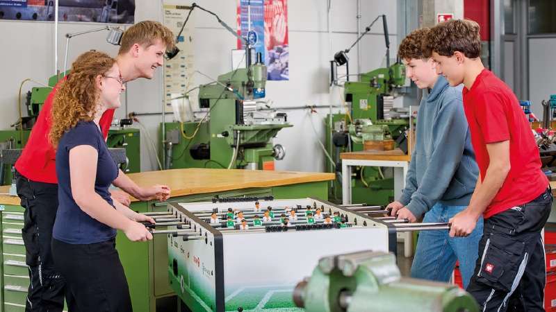 Quatre jeunes, deux de chaque côté, jouent au baby-foot dans un atelier industriel. Ils ont le sourire aux lèvres et sont concentrés, avec des machines vertes en arrière-plan.