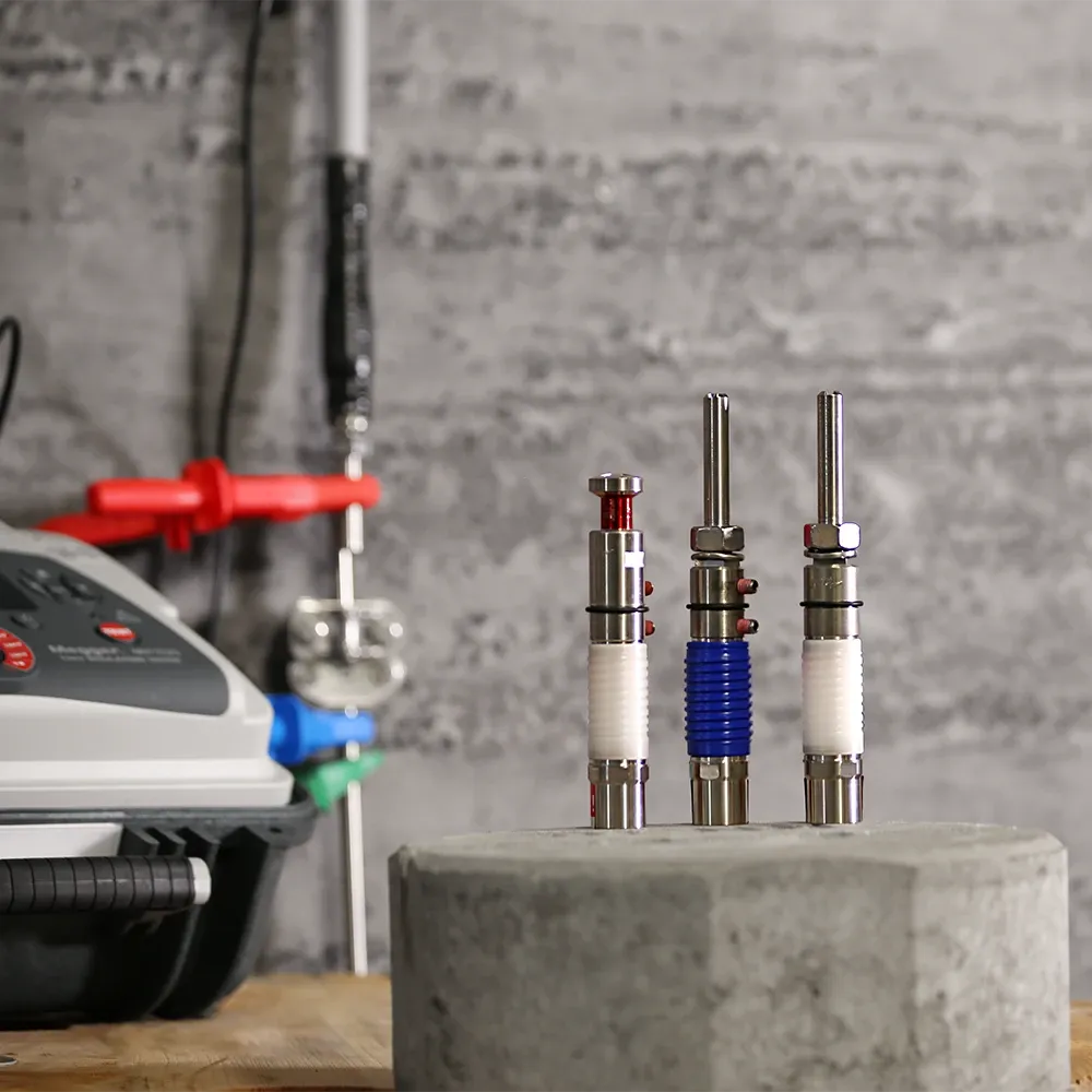 Trois objets cylindriques en métal, avec des composants rouges, bleus et blancs, sur une surface en béton devant un appareil équipé d’un panneau de commande. 