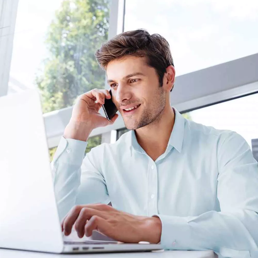 Man in a light blue shirt talking on a smartphone while working on a laptop in a bright office setting.