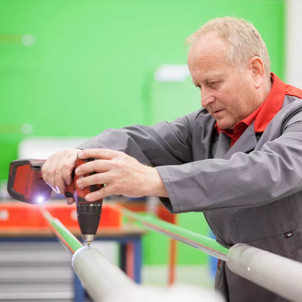 Worker in a workshop using a cordless drill to fasten a metal rod onto a cylindrical component.
