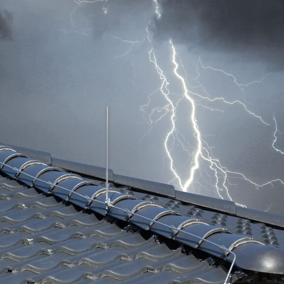 Rooftop lightning protection with metal conductors and air terminals on dark roof tiles, with lightning in a stormy sky behind.