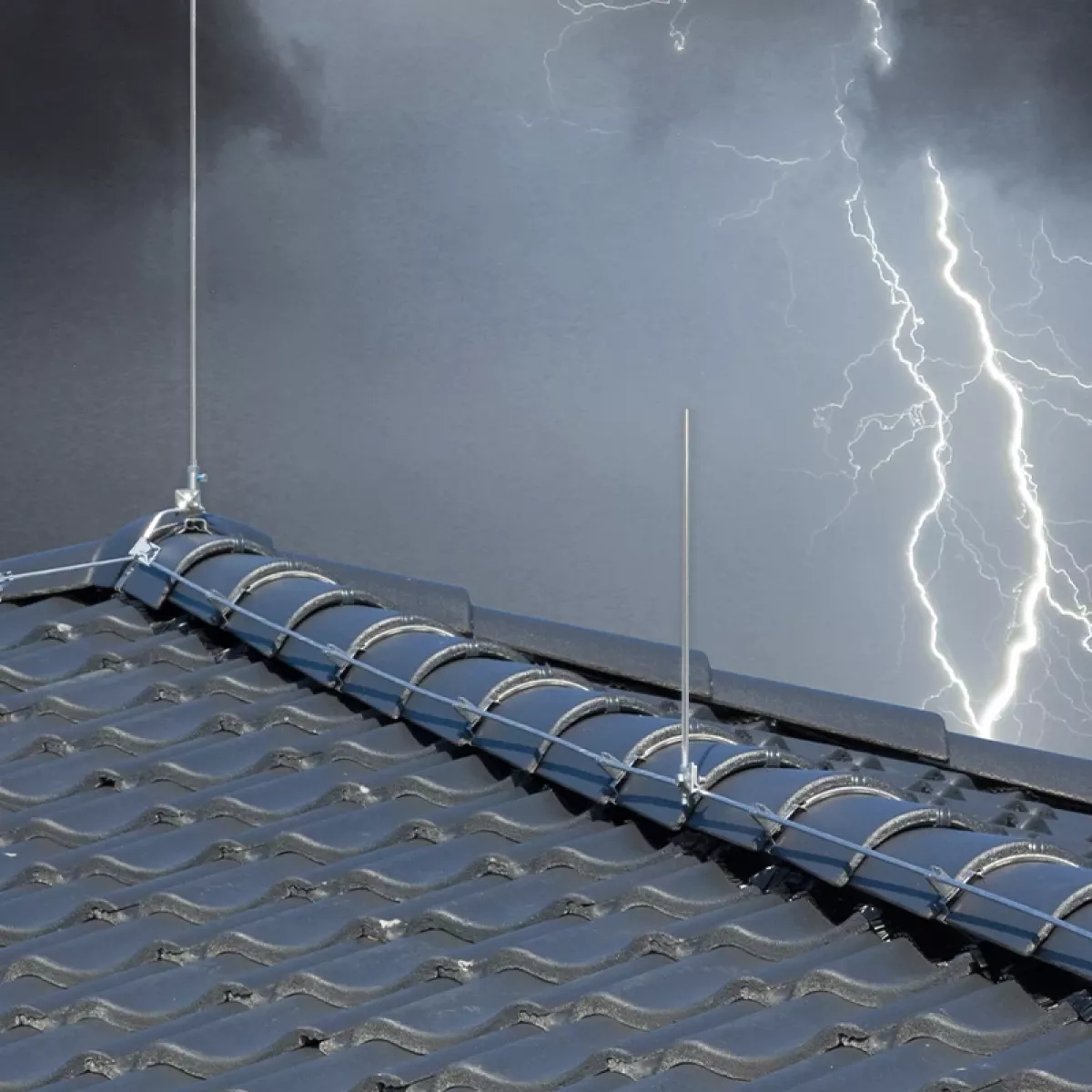 Rooftop lightning protection with metal conductors and air terminals on dark roof tiles, with lightning in a stormy sky behind.