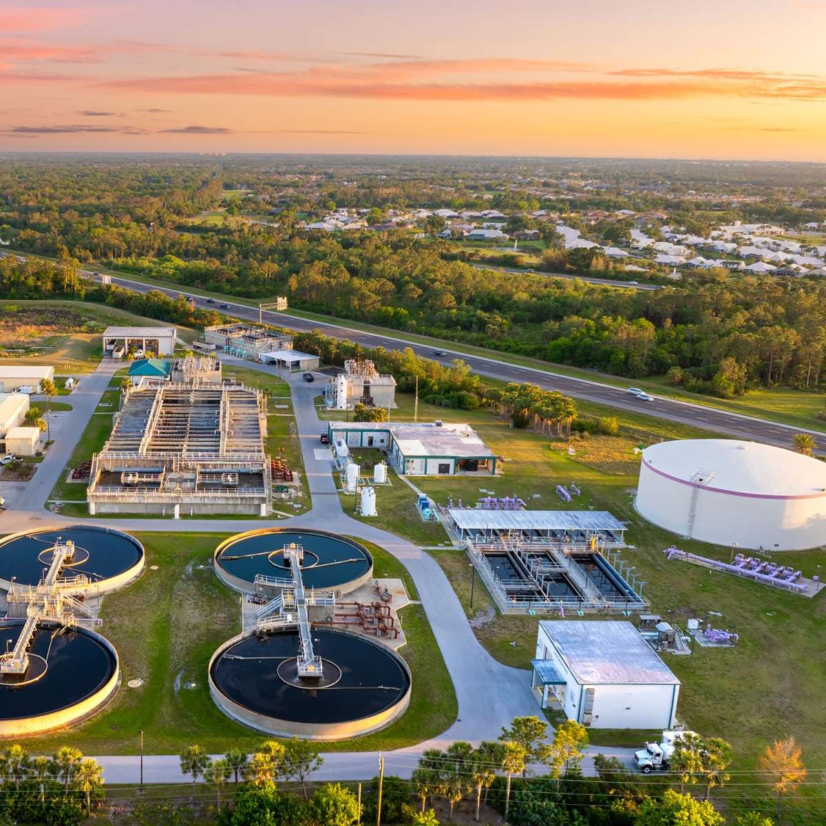 Aerial view of a water treatment facility with circular tanks, processing structures, roads, and surrounding greenery.