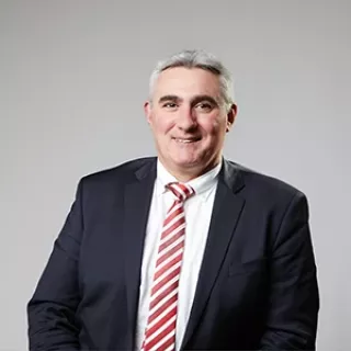Portrait of a man in a dark suit, white shirt, and red striped tie, smiling against a plain light background.