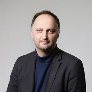 Portrait of a man with short hair wearing a dark blazer and navy top, facing the camera against a plain light background.