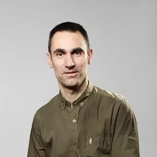 A studio portrait of a man with short dark hair, wearing an olive-green buttoned shirt.