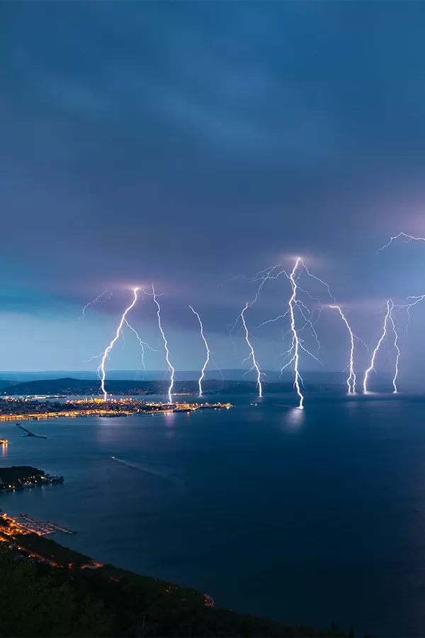 Multiple lightning bolts striking over a large body of water near a coastal city at night