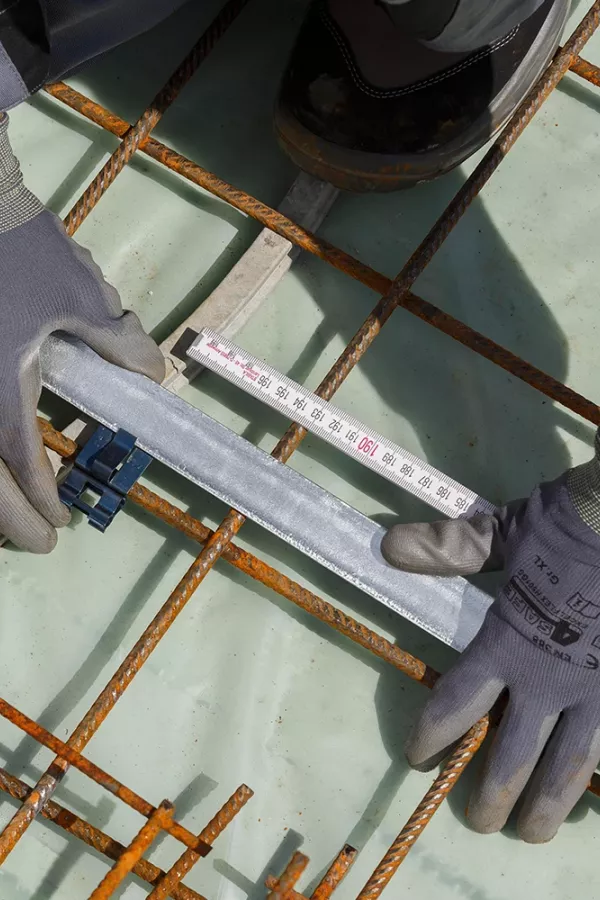 Gloved hands measure a flat metal strip on a steel rebar grid using a measuring tape with visible centimeter markings.