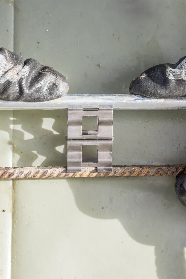 Gloved hands pressing a metal clip between two parallel steel rods against a light-colored surface.