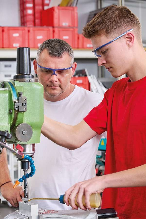 Deux hommes portant des lunettes de sécurité travaillent avec une perceuse à colonne verte dans un atelier. L'un d'eux fait fonctionner la machine, tandis que l'autre supervise, l'air très concentré.