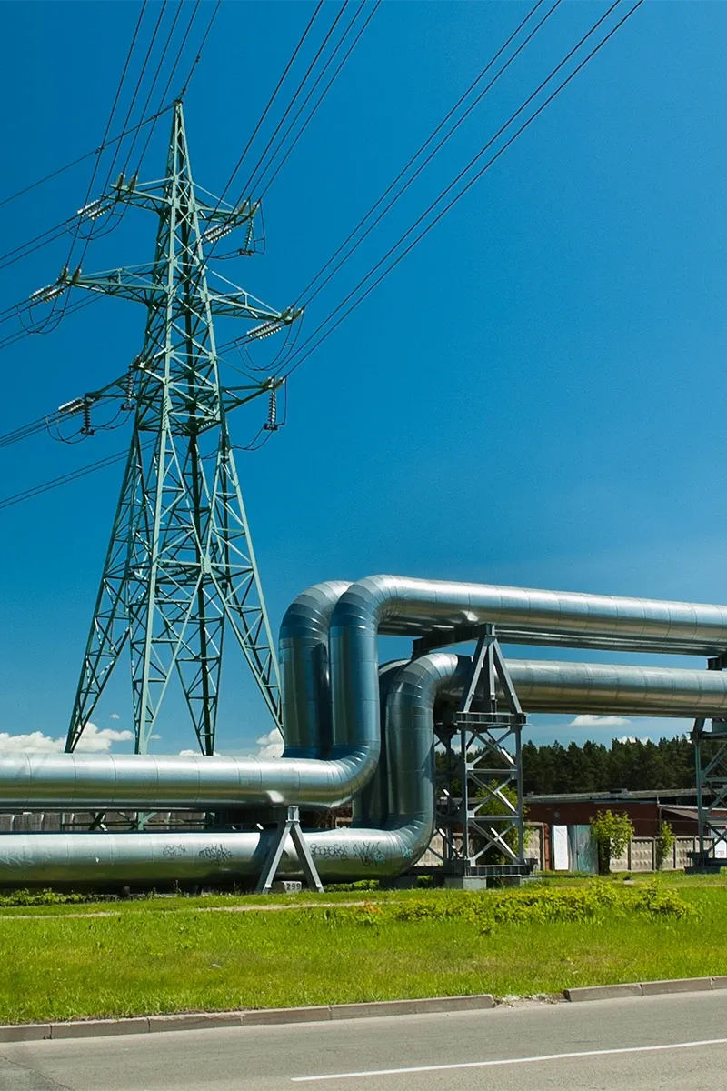 Grand pylône électrique vert derrière un pipeline, sur fond de prairie sous un ciel bleu.