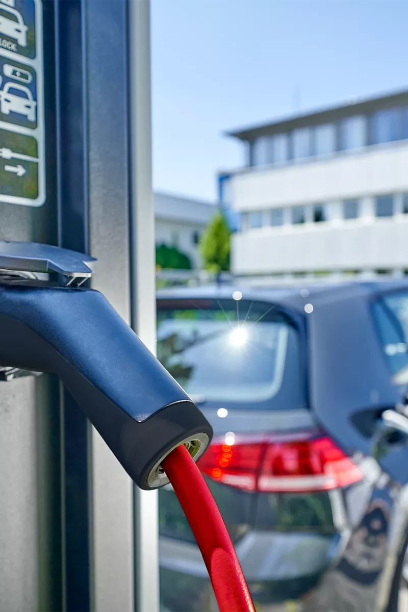 Close-up of an electric vehicle charging connector with red cable plugged into a charging station, car in background.