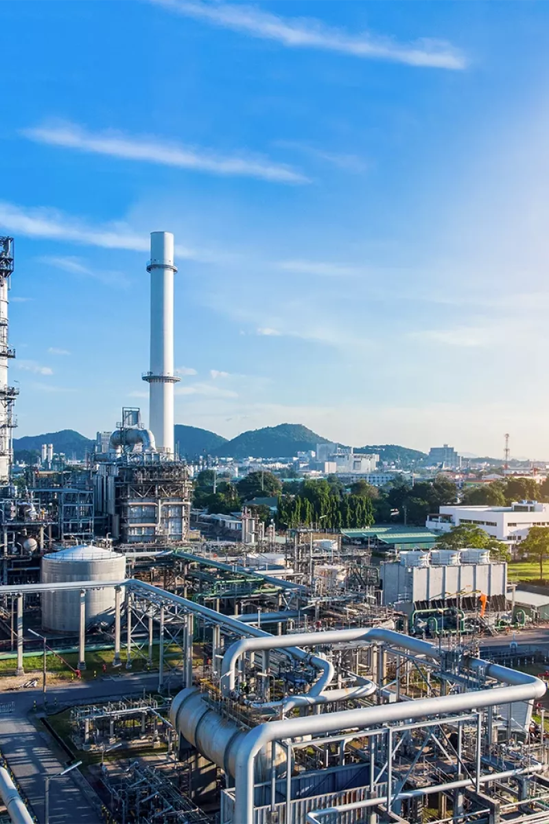 Industrial complex with pipes, storage tanks, and tall chimneys under a blue sky; mountains and a cityscape are visible in the background.