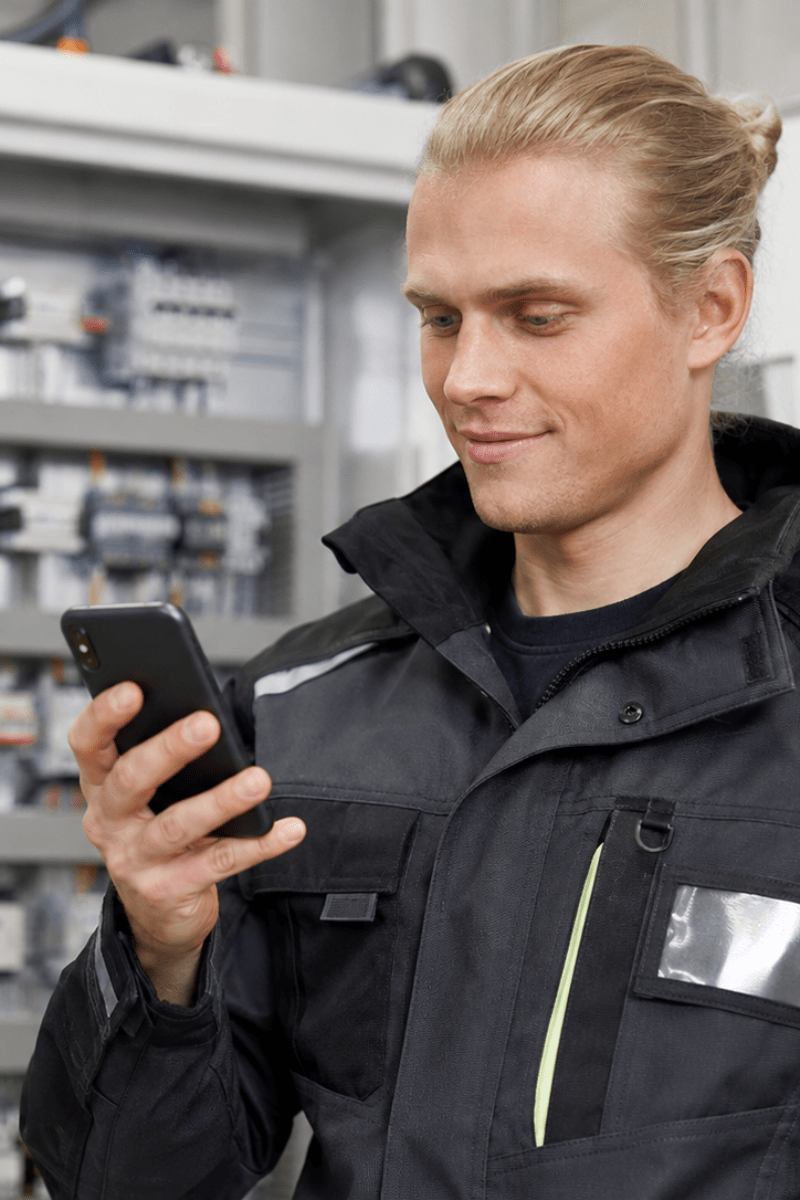 Photo of a man with long blond hairs in a ponytail and an electrical installer jacket front of an electrical panel with a smartphone
