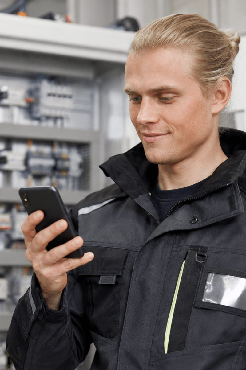 Photo of a man with long blond hairs in a ponytail and an electrical installer jacket front of an electrical panel with a smartphone