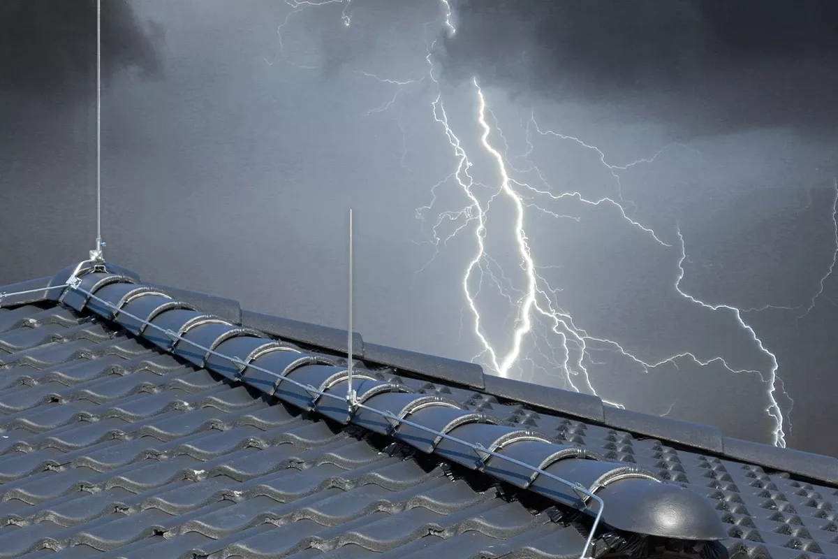 Rooftop lightning protection with metal conductors and air terminals on dark roof tiles, with lightning in a stormy sky behind.