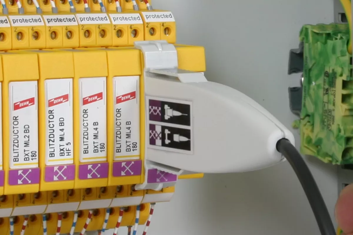 White test adapter plugged into a yellow surge protection module inside an electrical control cabinet with connected wires.