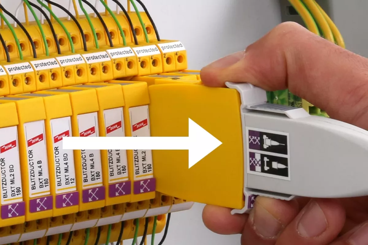 Hand removing a yellow surge protection module from a control cabinet while holding a white test adapter; white arrow indicates direction.