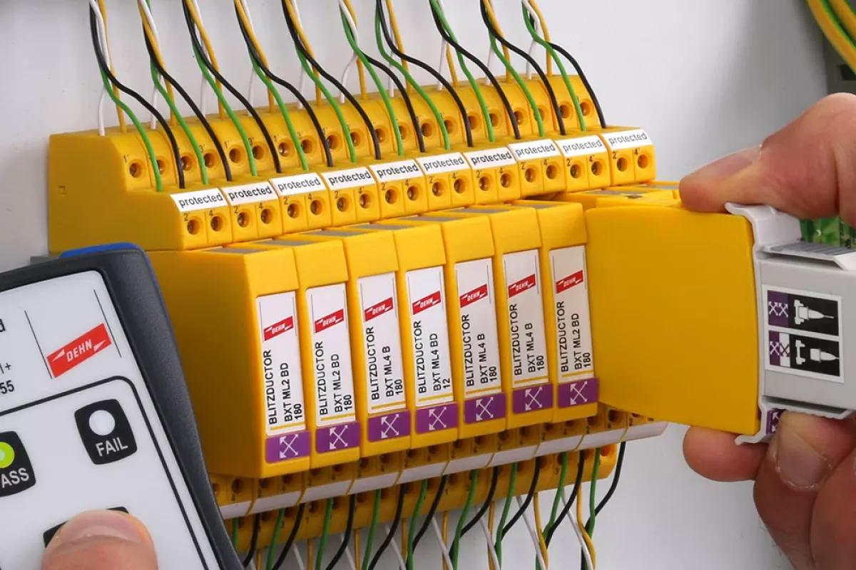 Hand holding a measuring device next to a row of yellow surge protection modules while another hand holds a removed module with test adapter.