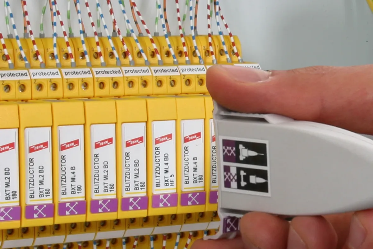 Hand holding a white test adapter next to yellow surge protection modules inside a control cabinet with connected wires.