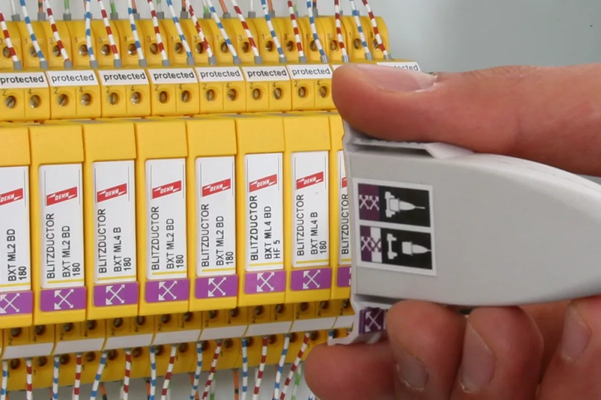 Hand holding a white test adapter next to yellow surge protection modules inside a control cabinet with connected wires.